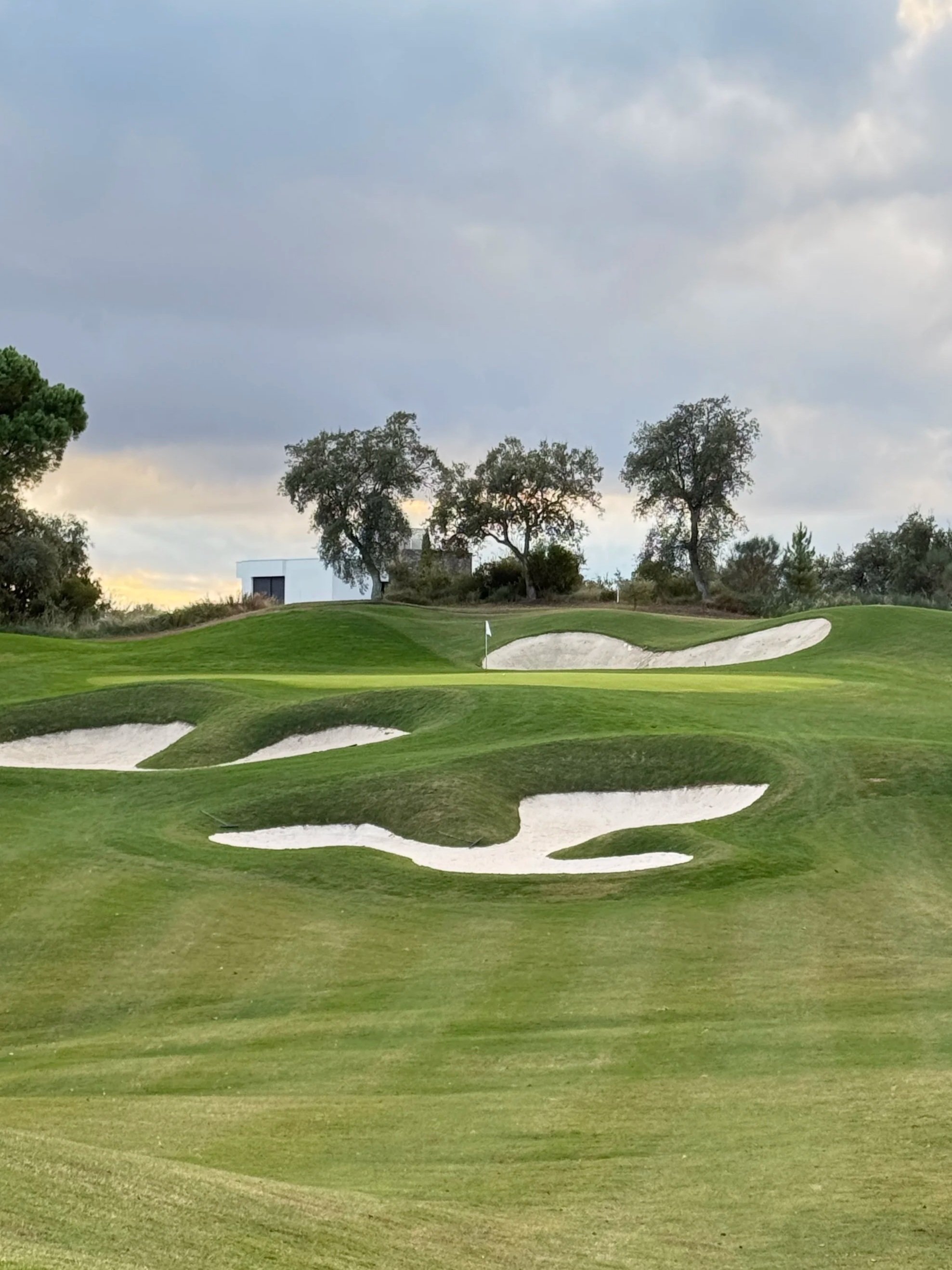 A golf course landscape with a green, multiple sand bunkers, and a flagstick. Trees and a modern building are in the background, under a partly cloudy sky.