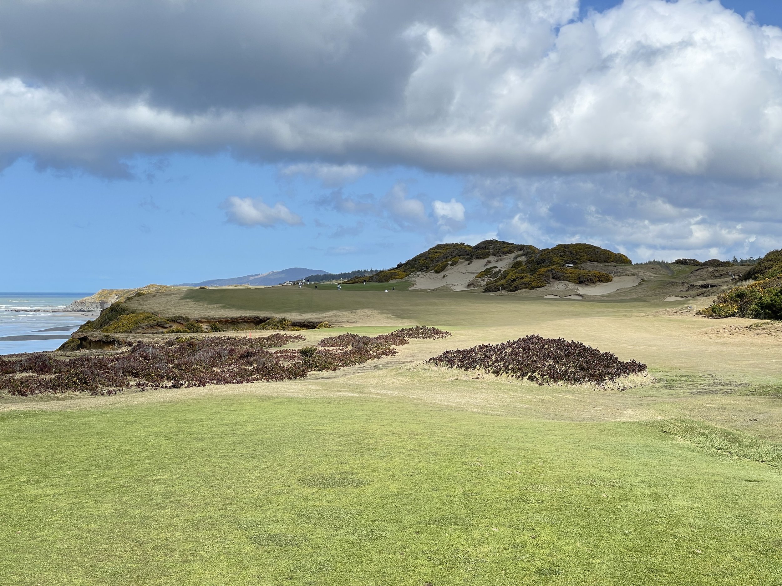 A scenic view of a golf course near the coast with green grass, bushes, and rolling hills under a partly cloudy sky.