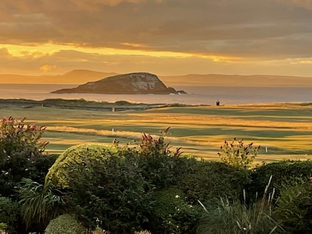 Scenic view of a golf course near the coast at sunset, with a large island or hill in the background and lush green vegetation in the foreground.