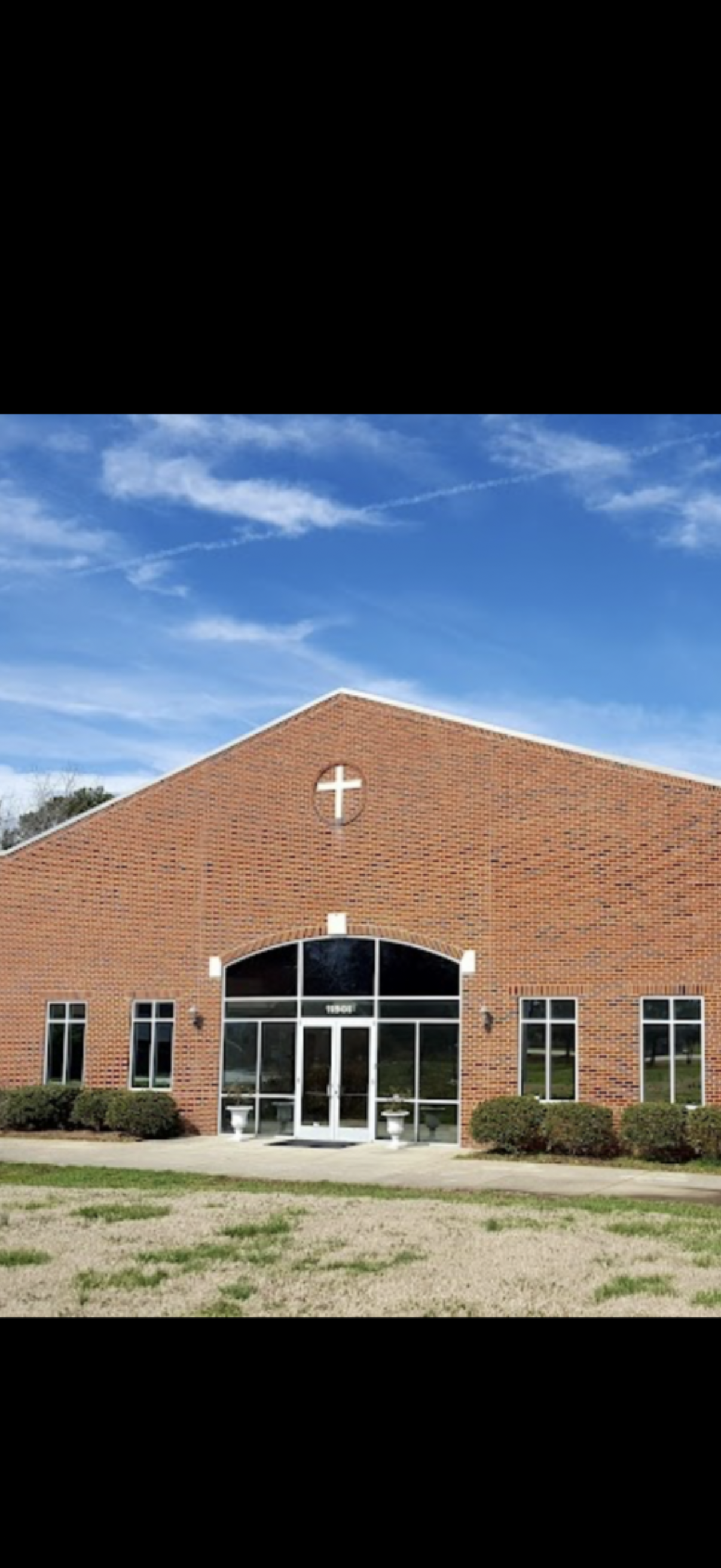 A brick church building with a large cross on the circle window near the roof, multiple windows, and a white door, with a blue sky in the background.