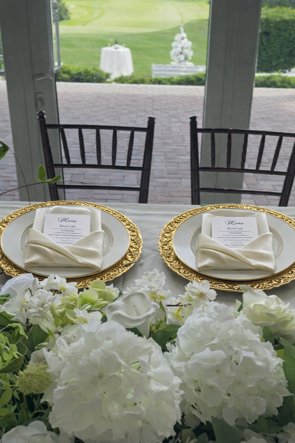 Elegant table setting with two plates, each with a folded napkin and menu, surrounded by white flowers, with a patio and garden in the background.