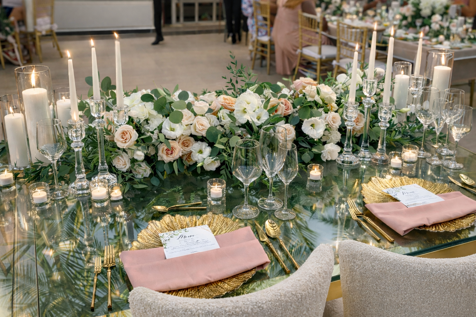 Elegant wedding table with floral centerpiece, candles, glassware, gold-rimmed plates, pink napkins, and gold utensils.