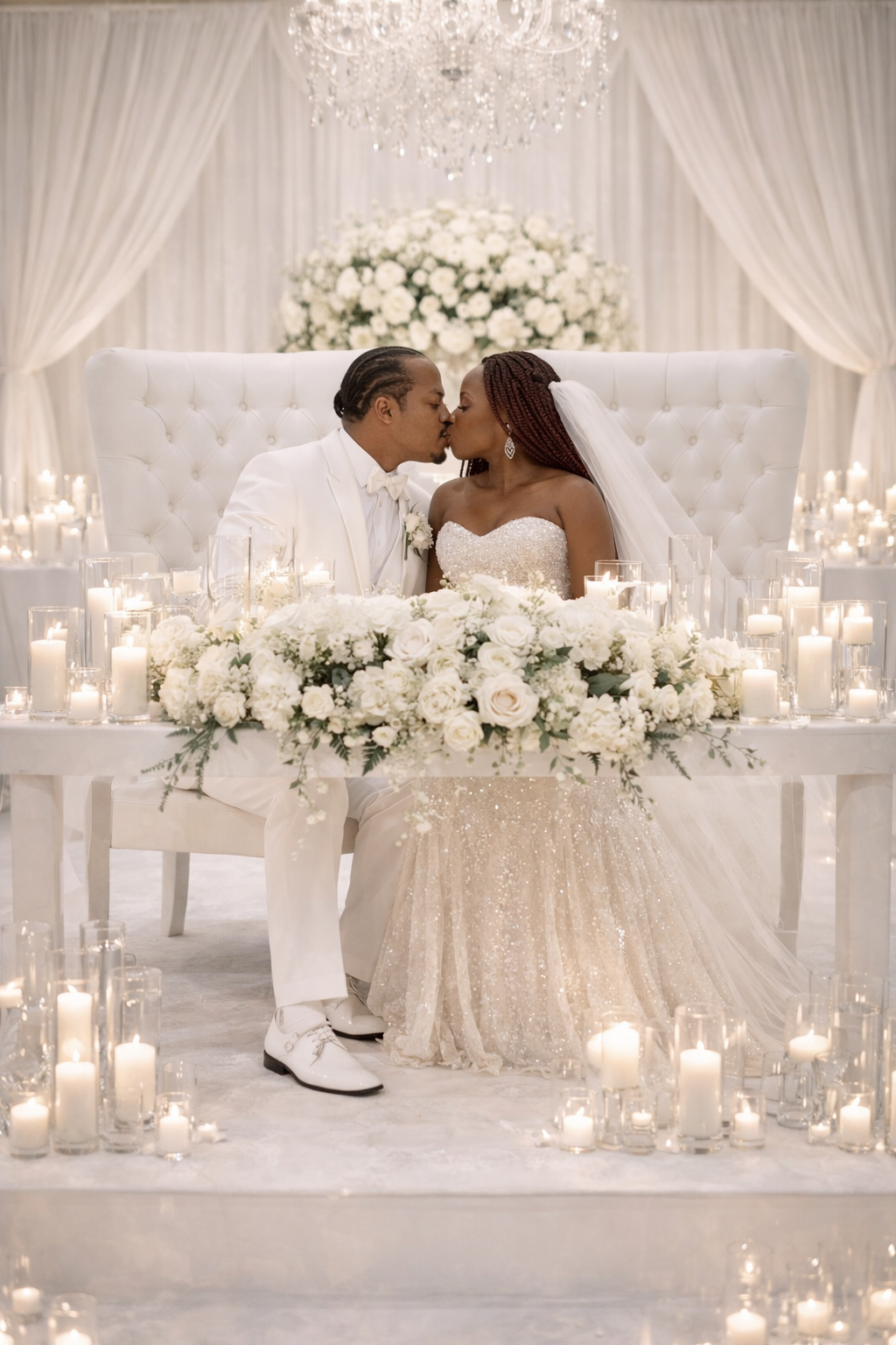 Elegant luxury wedding reception featuring bride and groom seated at a candlelit sweetheart table with white floral arrangements, crystal chandelier, and romantic all-white decor by ReneJohn Designs.