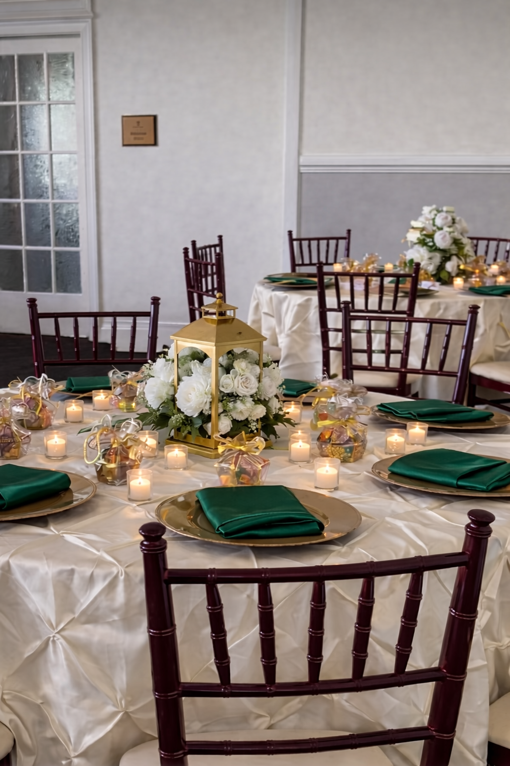 Round banquet table decorated with white tablecloth, green napkins, gold chargers, and candlelit centerpieces. A gold lantern with white roses and greenery serves as the main centerpiece, surrounded by small tea light candles and gift bags with ribbons. Multiple chairs are arranged around the table, with additional tables and chairs in the background.