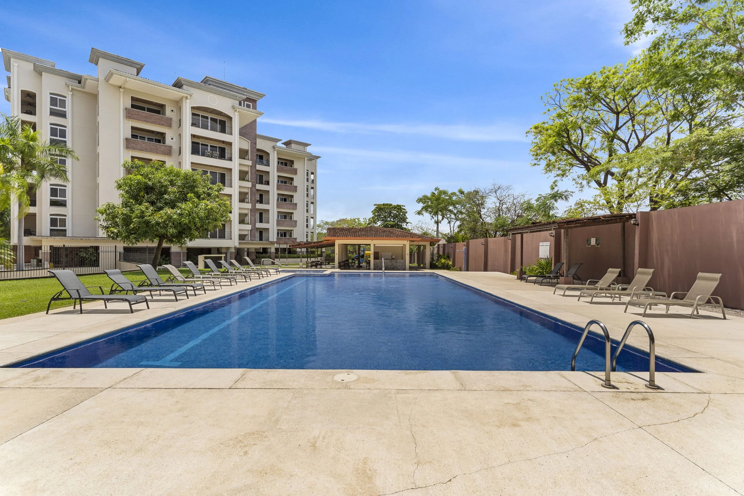 Outdoor swimming pool with lounge chairs along the sides, surrounded by trees and a multi-story residential building in the background, under a bright blue sky.