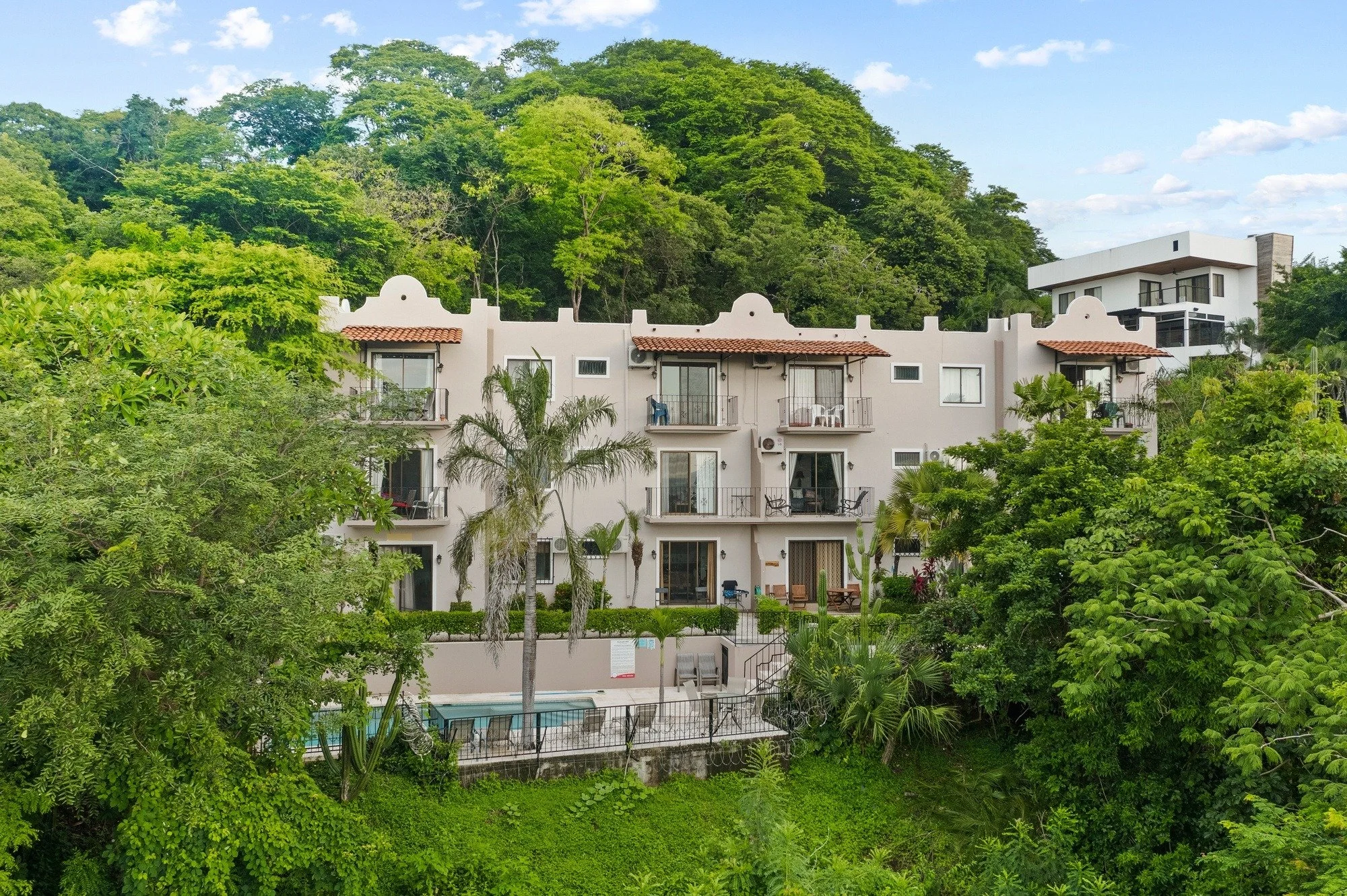 White multi-story apartments with balconies, surrounded by green trees, with a hill in the background and a swimming pool in front.