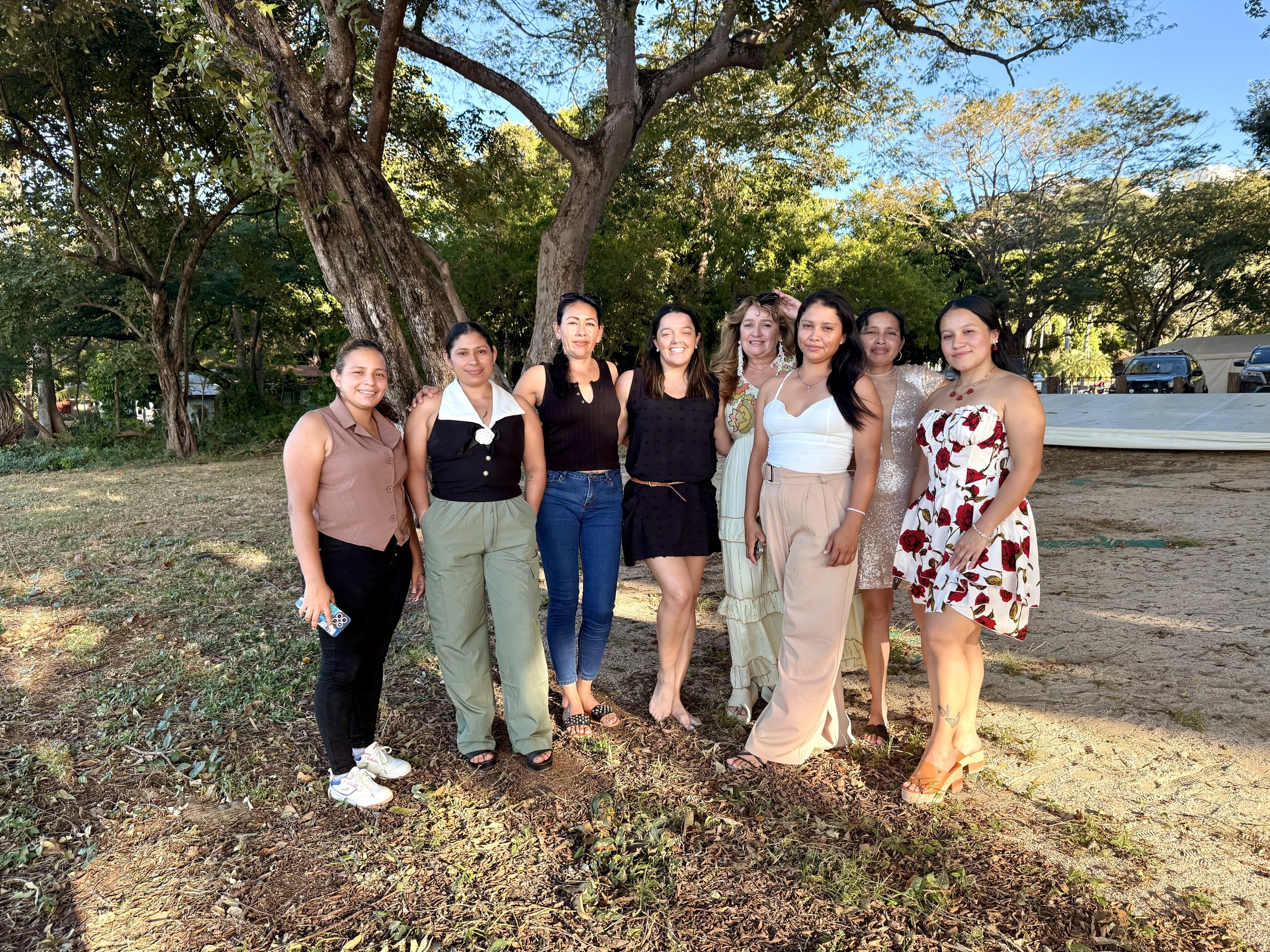 A group of nine women posing outdoors with trees and a clear blue sky in the background.