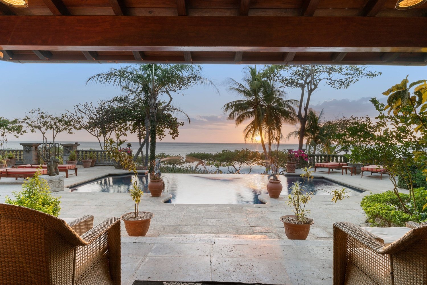 A patio view overlooking the ocean at sunset, featuring a pool, potted plants, lounge chairs, and palm trees.