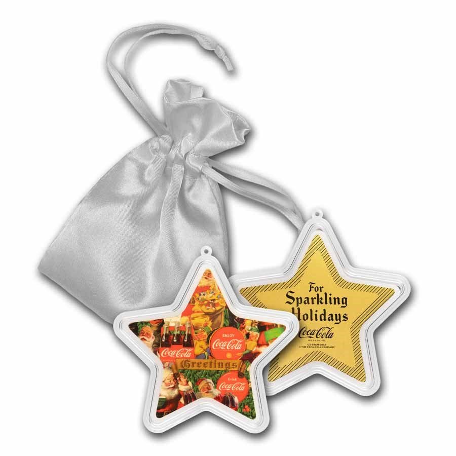 Holiday-themed Coca-Cola ornaments in star shapes, one with a festive scene and the other with a gold design, attached to a silver satin gift bag.