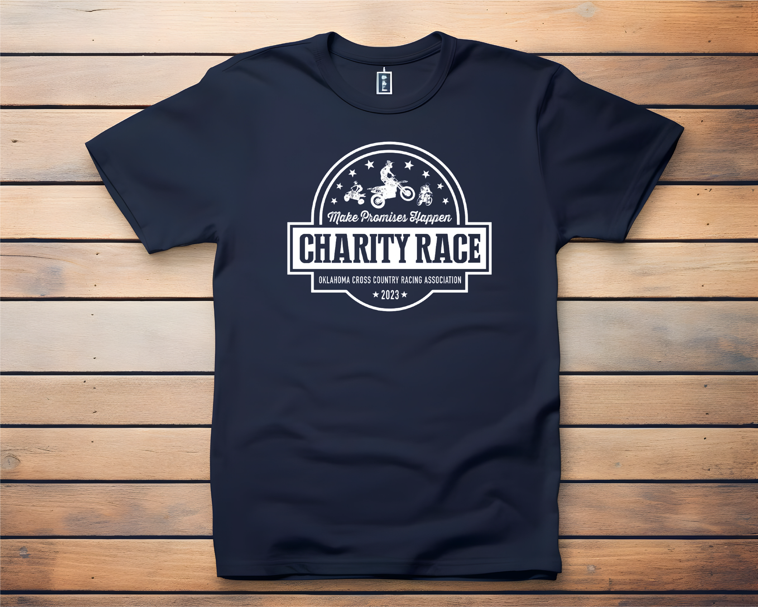 Navy blue T-shirt with a white graphic design promoting a charity race organized by the Oklahoma Cross Country Racing Association in 2023. The design features illustrations of children riding bicycles and a motorcycle, with stars and the phrase 'Make