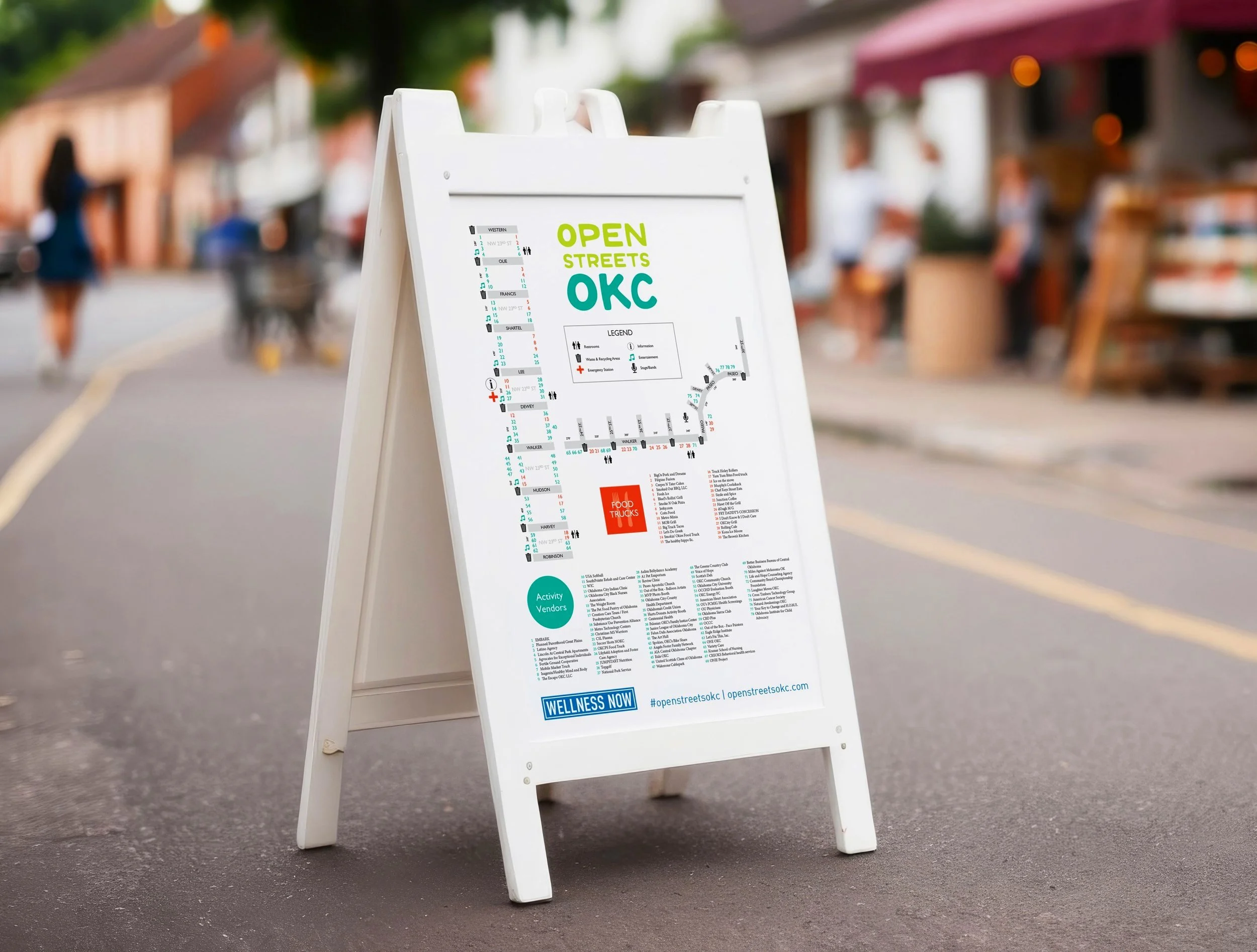A white A-frame signboard on a street displaying a map and information for the Open Streets OKC event, with blurred people and shops in the background.