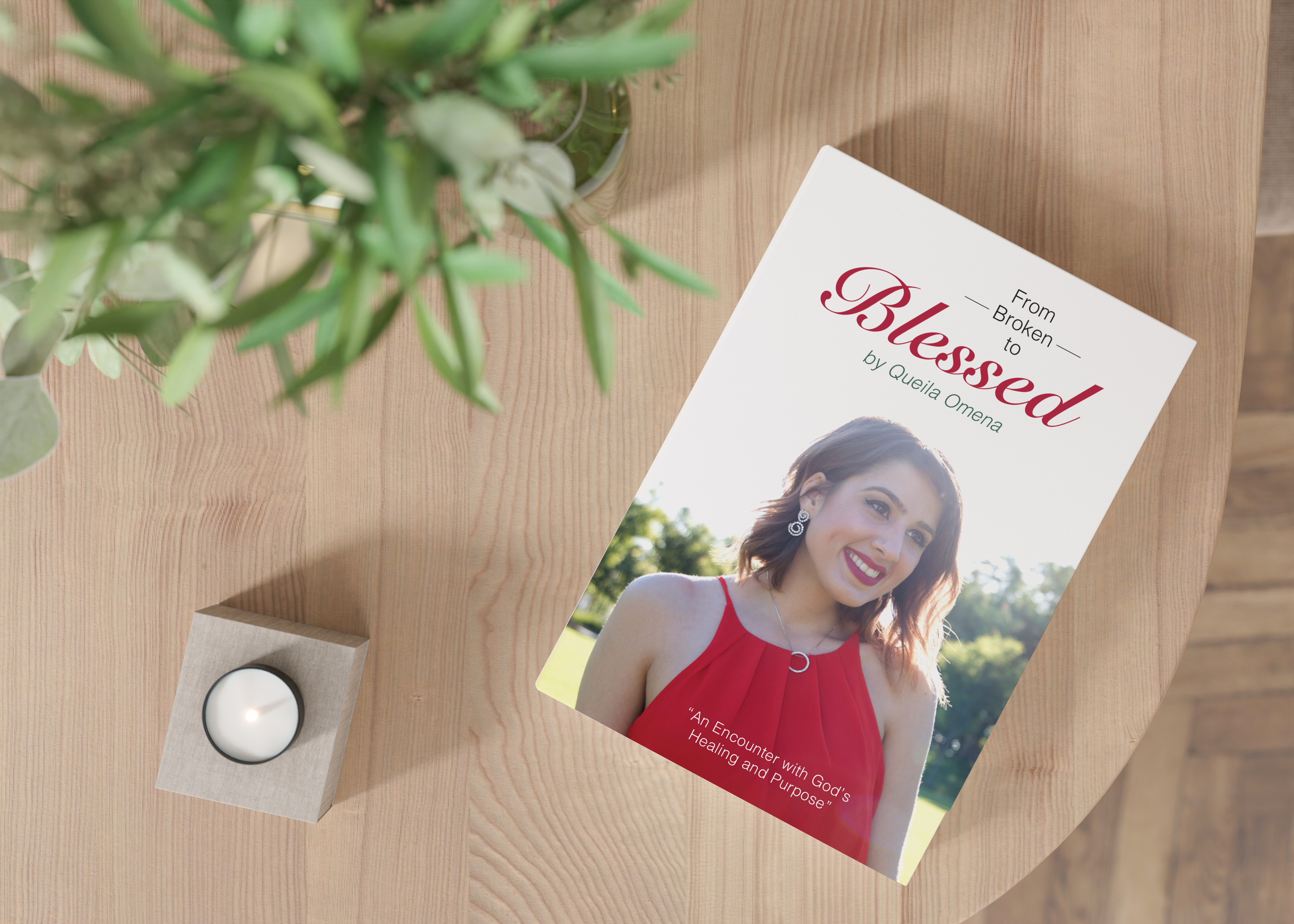A paperback book titled 'From Broken to Blessed' by Quella Omena is on a wooden table next to a small tealight candle and some green leaves.