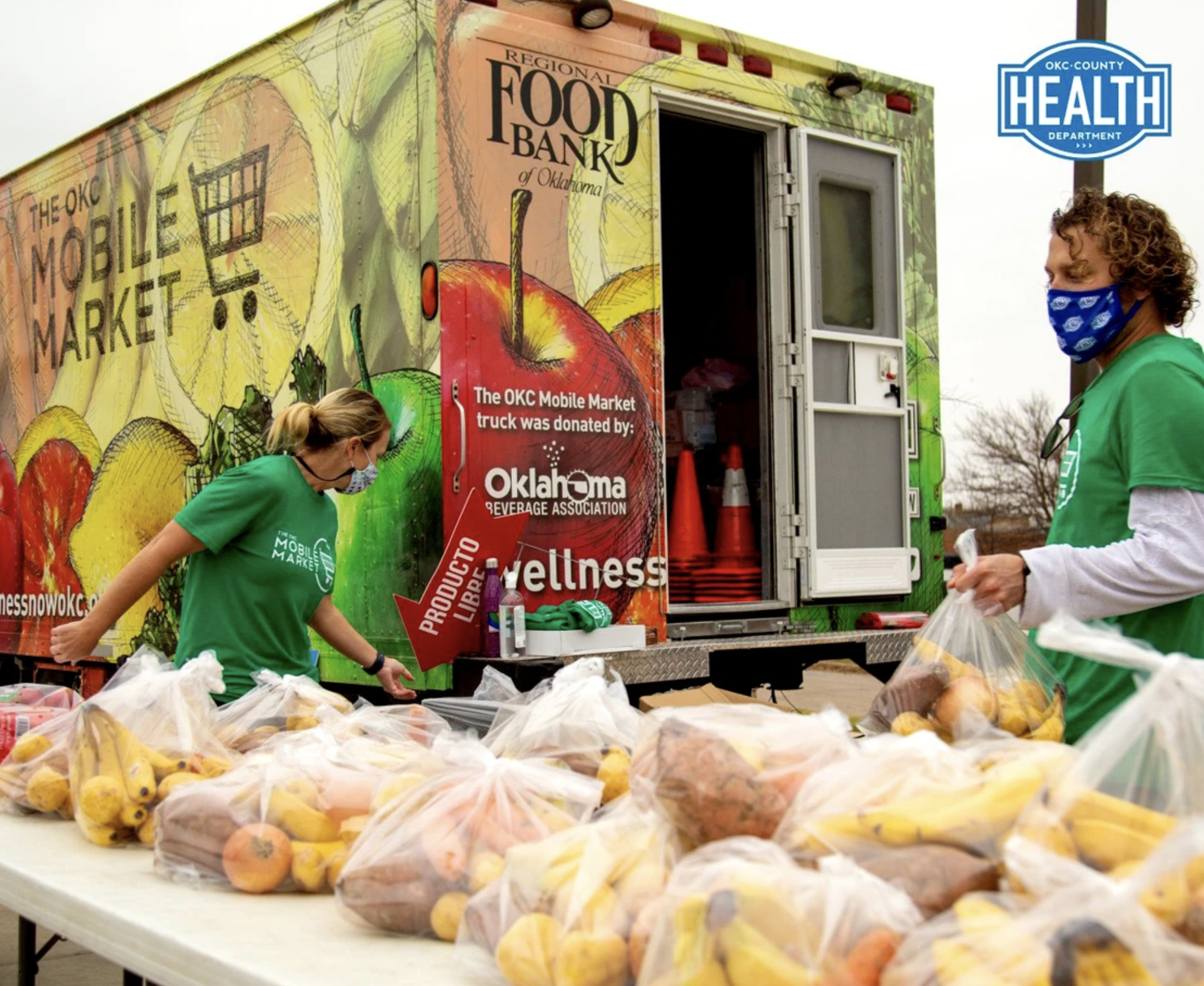 Two people wearing green t-shirts and face masks unloading produce from a mobile food bank truck with bags of fruit and vegetables on a table in front. The truck has colorful fruit artwork and signage indicating it's from the Oklahoma Food Bank and H