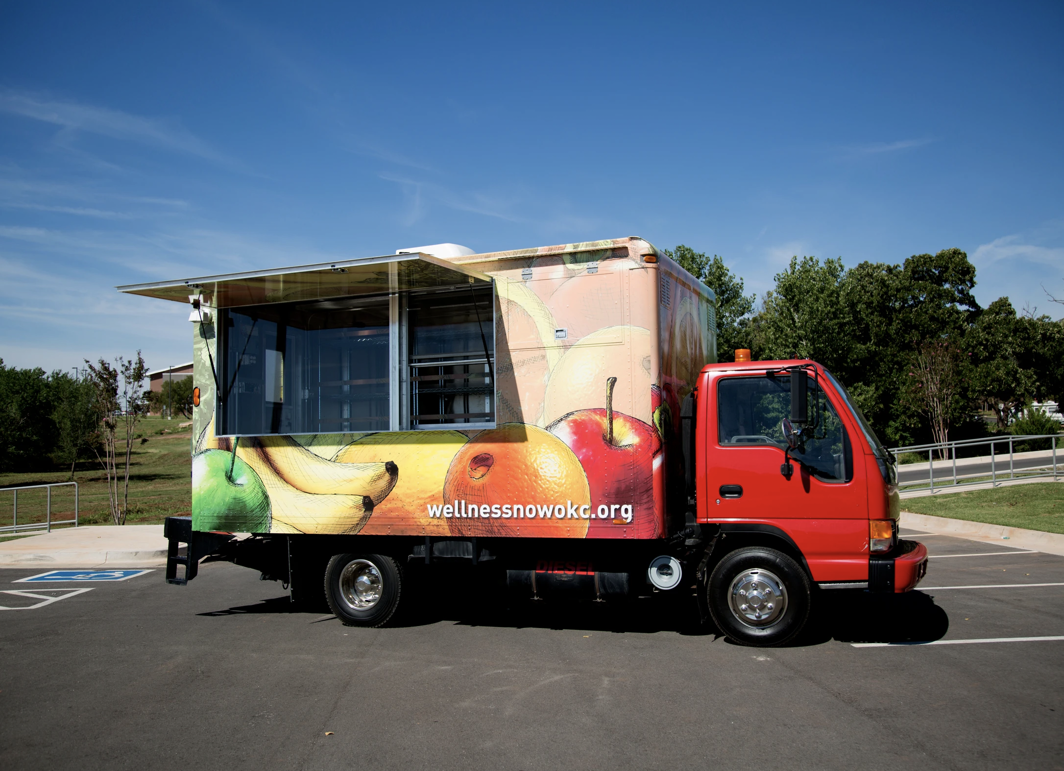 Food truck with fruit-themed wrap parked in a lot with trees and blue sky in the background.