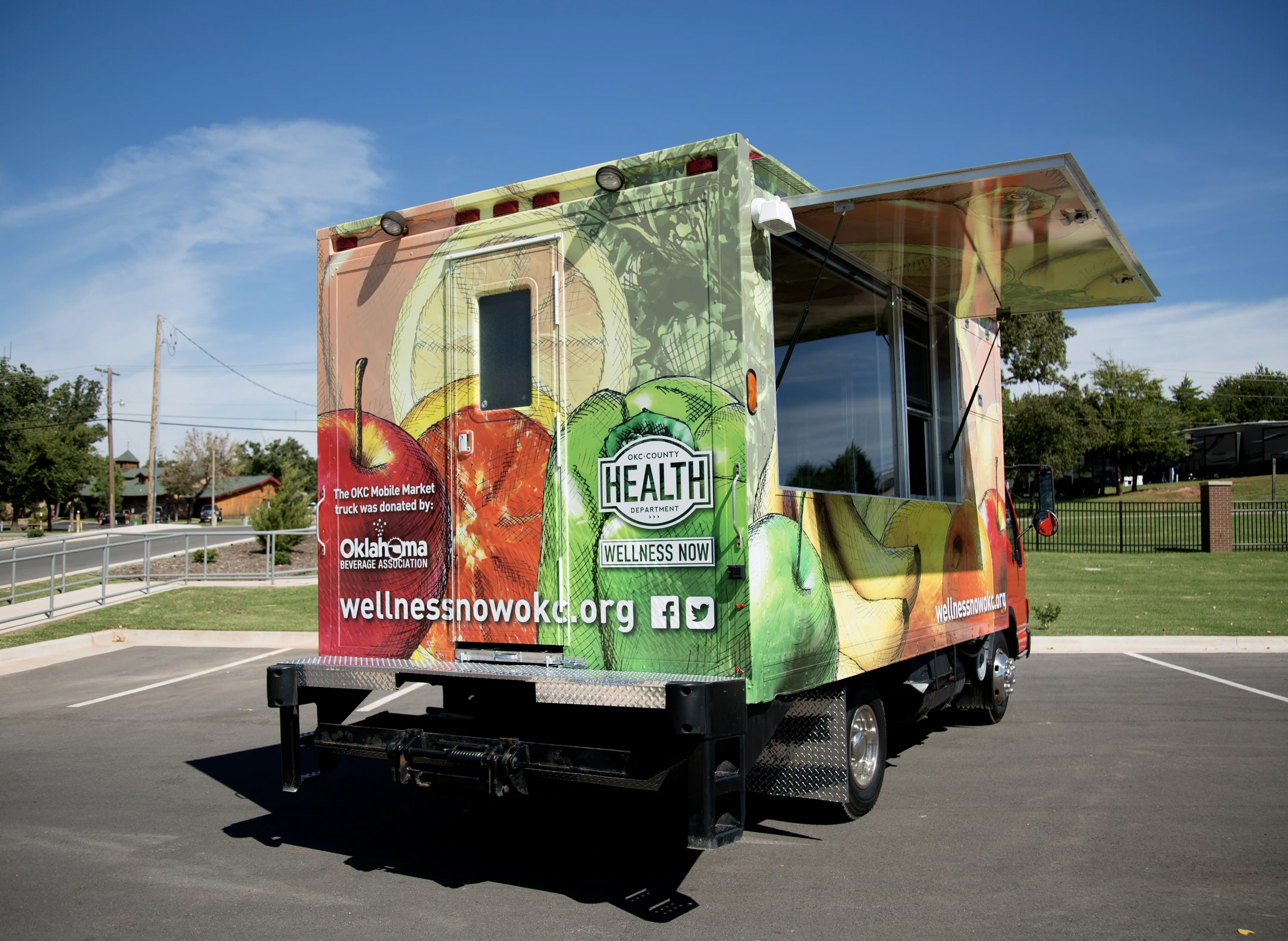 Mobile grocery truck covered in colorful fruit illustrations, parked in an empty lot with grass, trees, and a fence in the background.