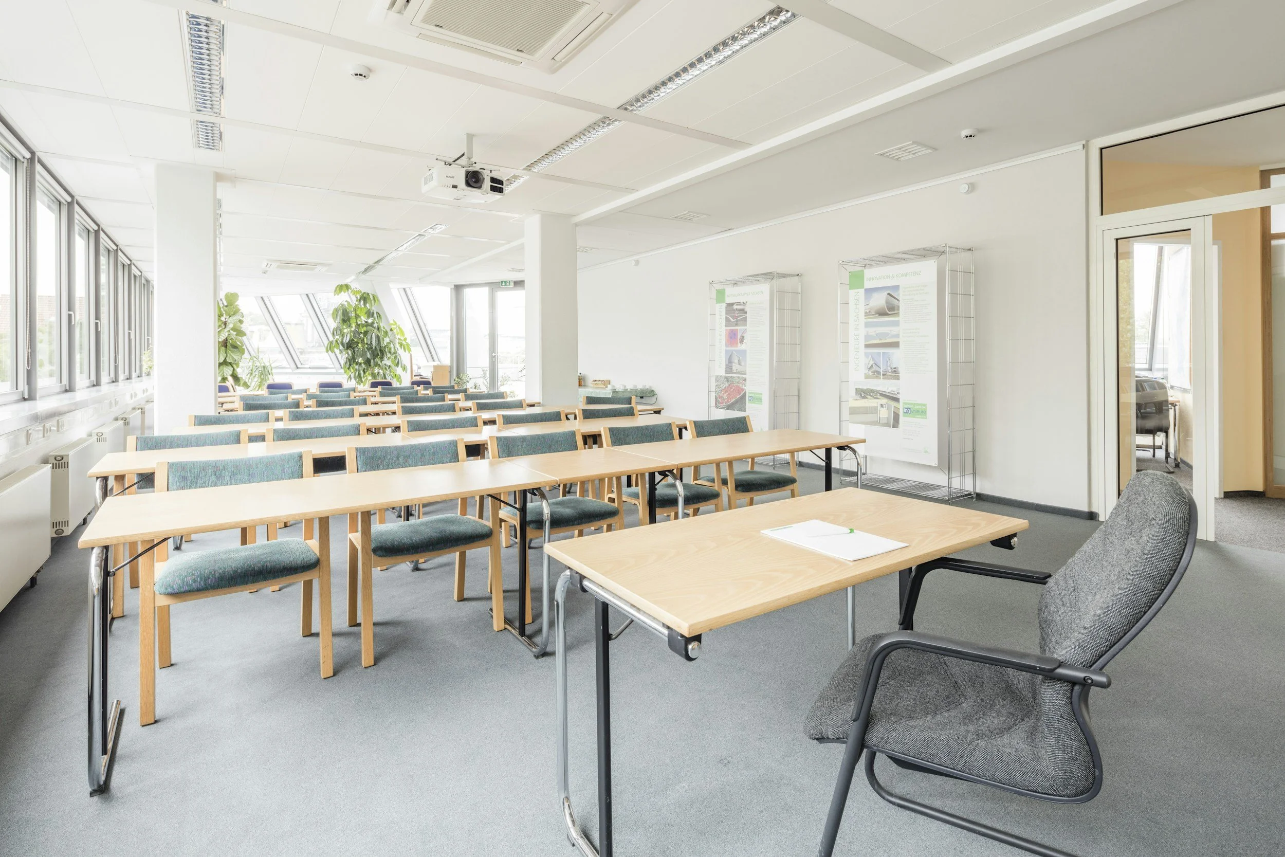 Empty conference room with long wooden tables, upholstered chairs, large windows, and display panels on the wall.