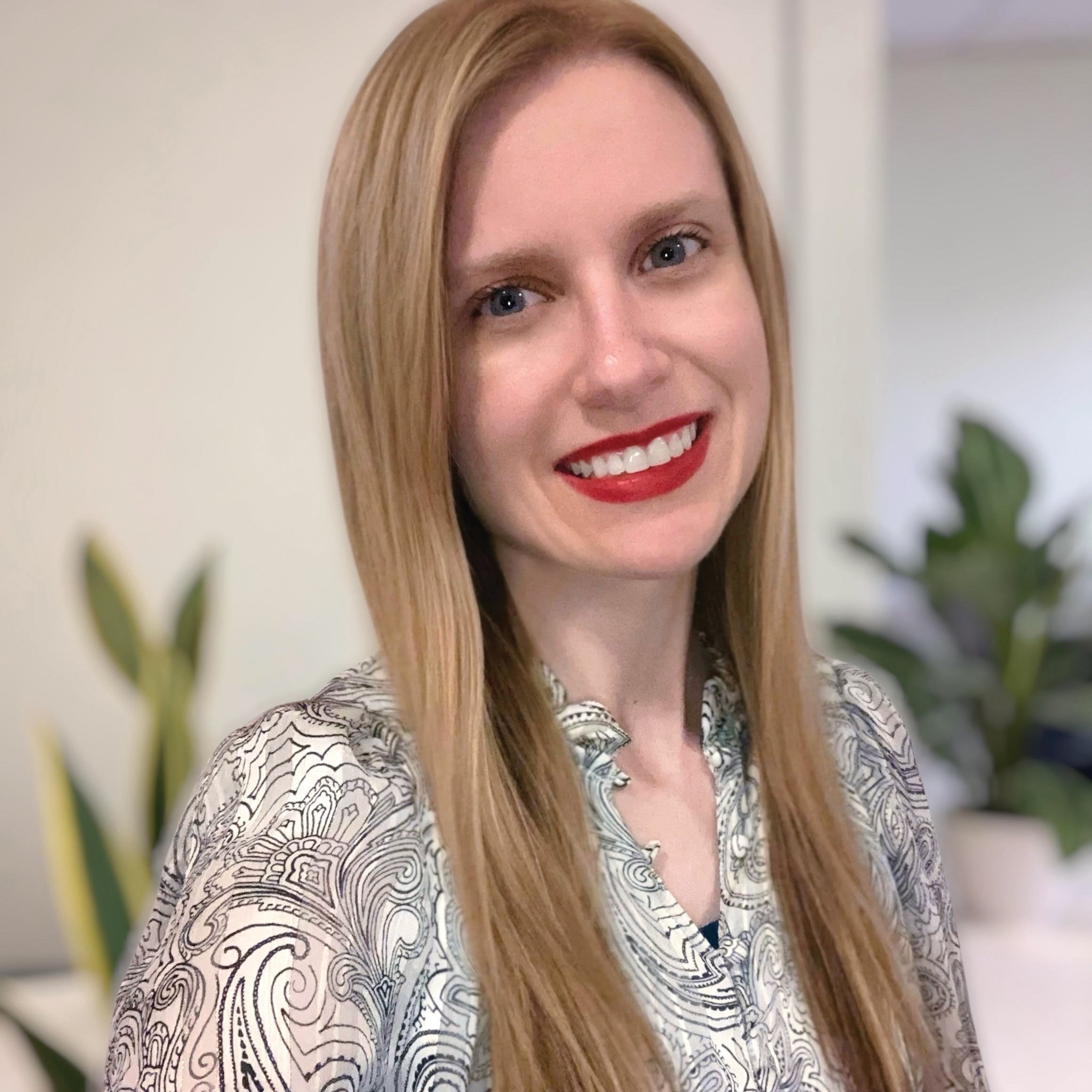 A woman with long, straight red hair, blue eyes, and red lipstick smiling, wearing a patterned blouse, with blurred green plants in the background.