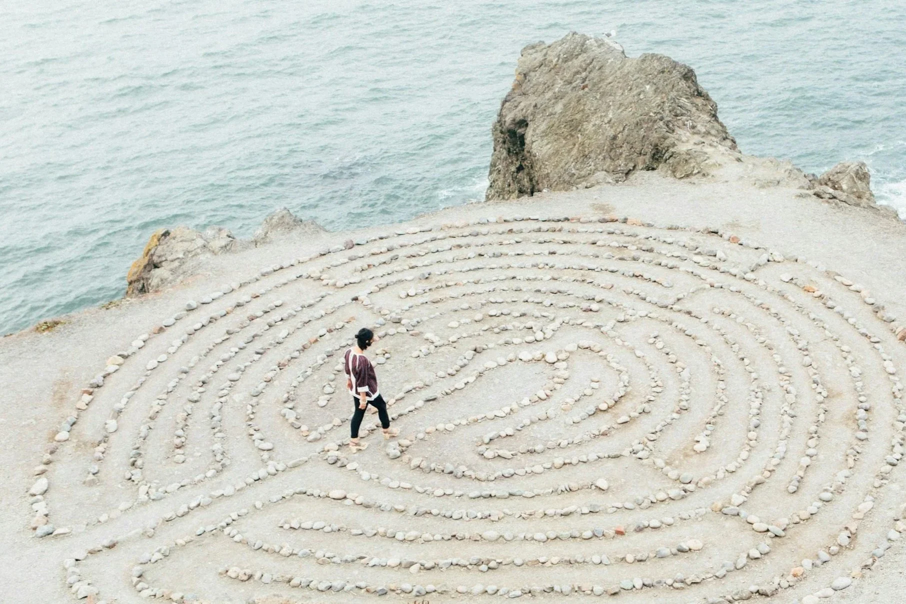 A person walking on a spiral rock labyrinth near the ocean, with large rocks and cliffs in the background.
