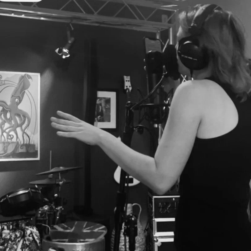 A woman recording vocals in a studio with headphones on, microphone, and music equipment, with artwork on the wall.