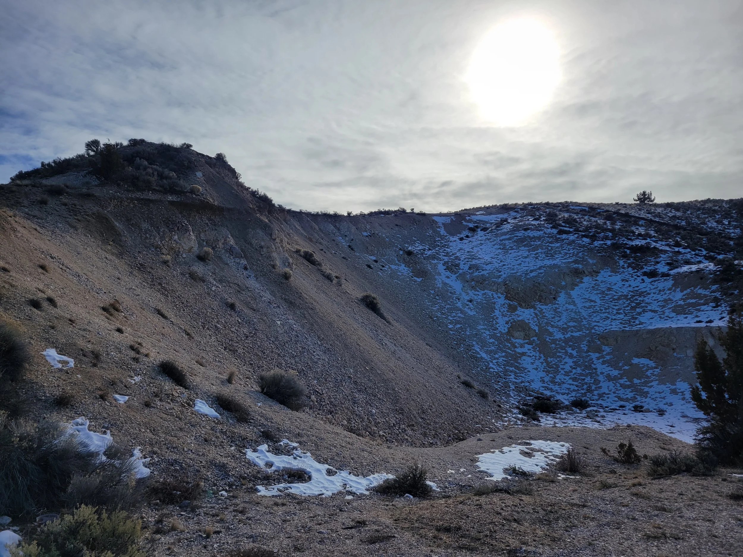 A rugged hillside with sparse vegetation and patches of snow, under a cloudy sky with the sun partially obscured.