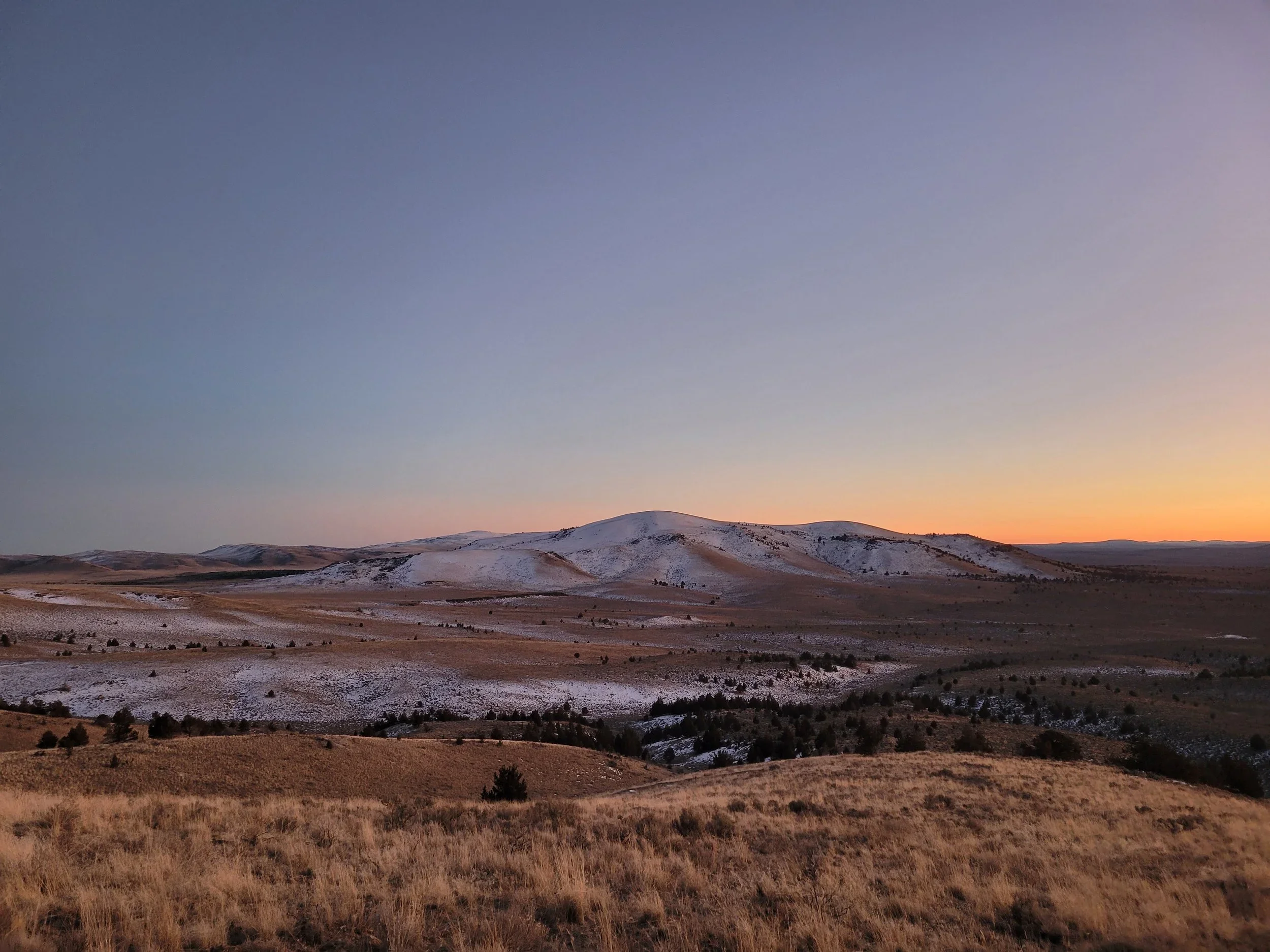 A landscape of rolling hills, some covered with snow, under a clear sky at sunset