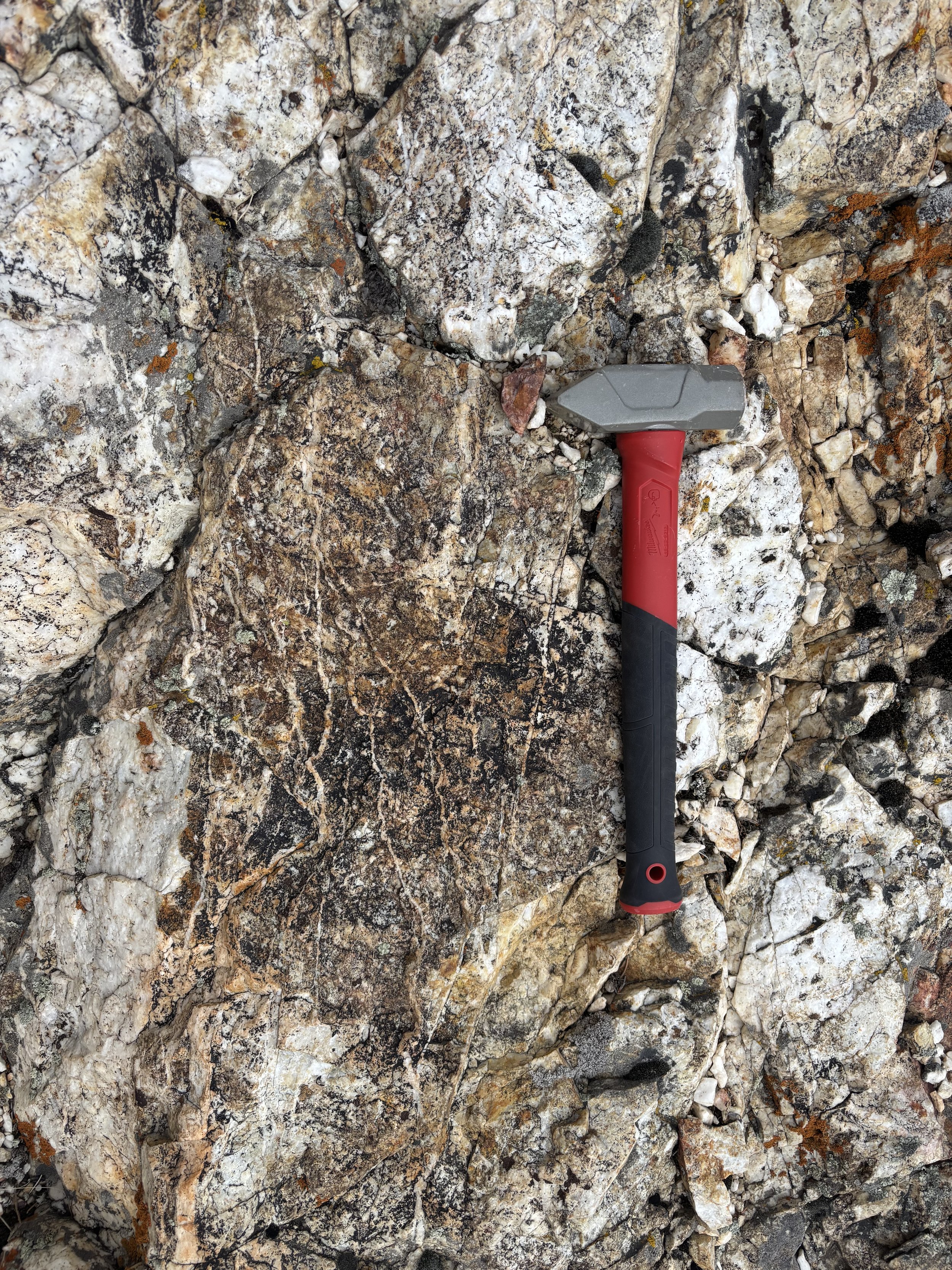 A hand-held hammer with a gray head and red and black handle lying on a bed of rocks and small stones.