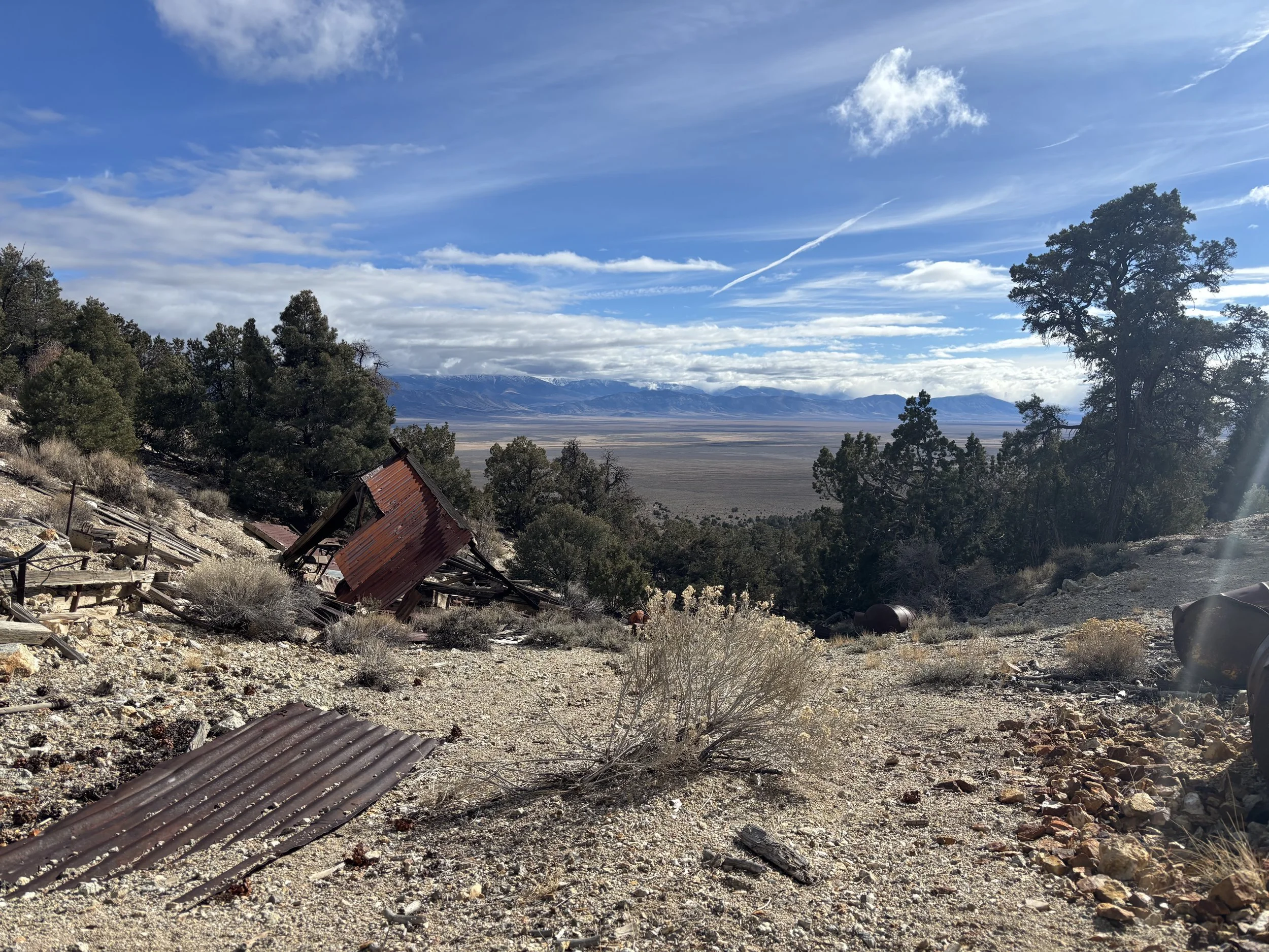 Scene showing a hillside with scattered debris and rusty metal sheets, sparse vegetation, and a mountain range in the distance under a partly cloudy sky.