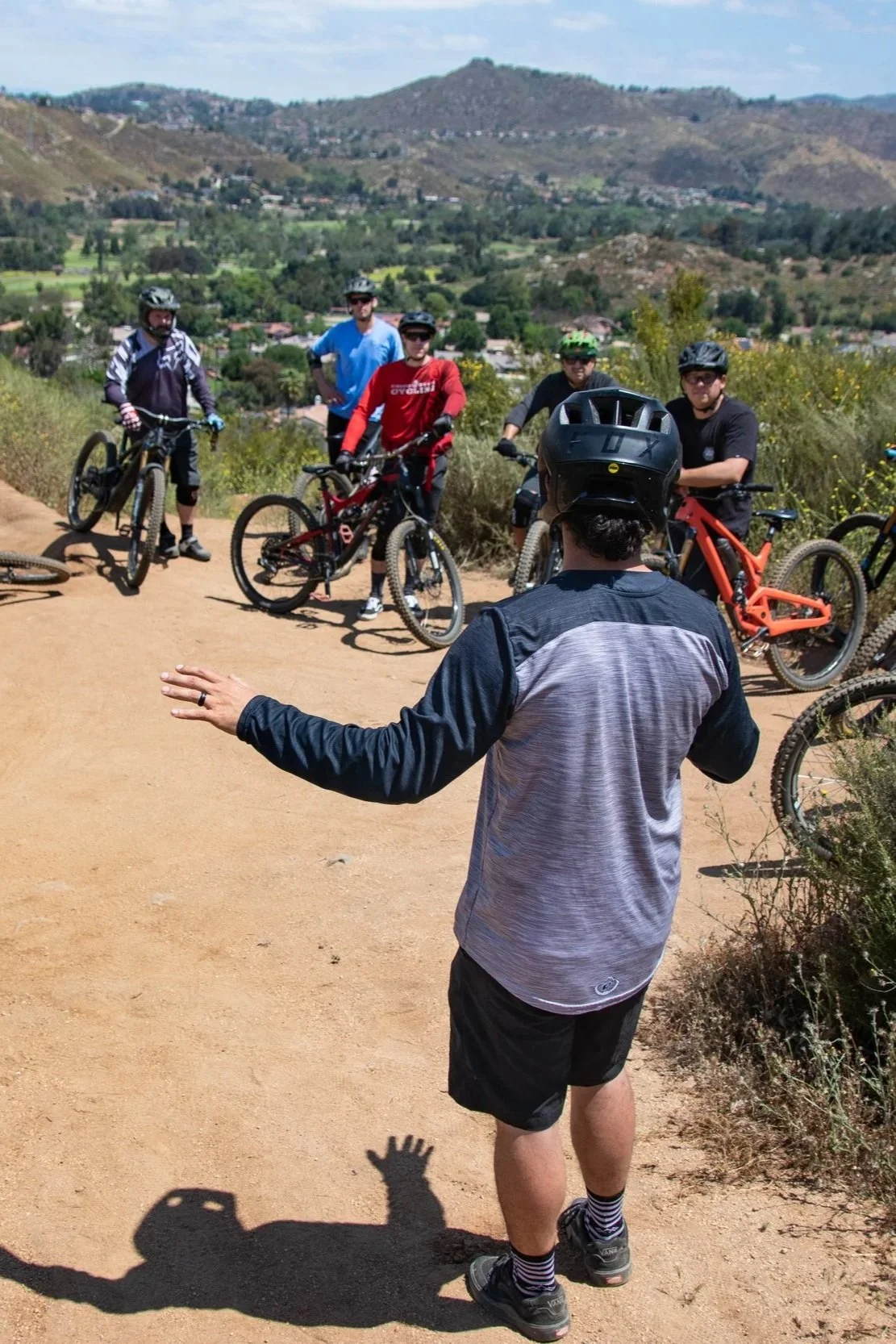 A group of five mountain bikers standing on a trail with a person in a helmet speaking to them. The background features a scenic view of hills, trees, and houses under a partly cloudy sky.