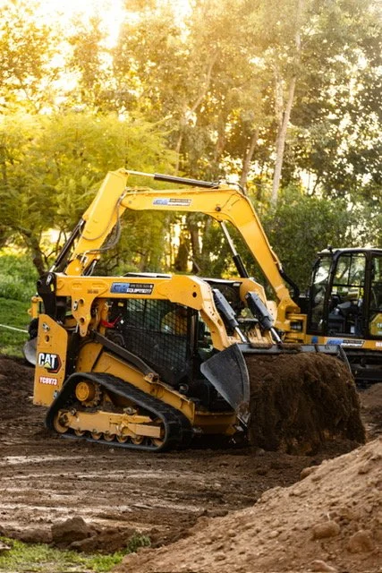 A yellow compact excavator digging soil in a construction site surrounded by trees at sunset.