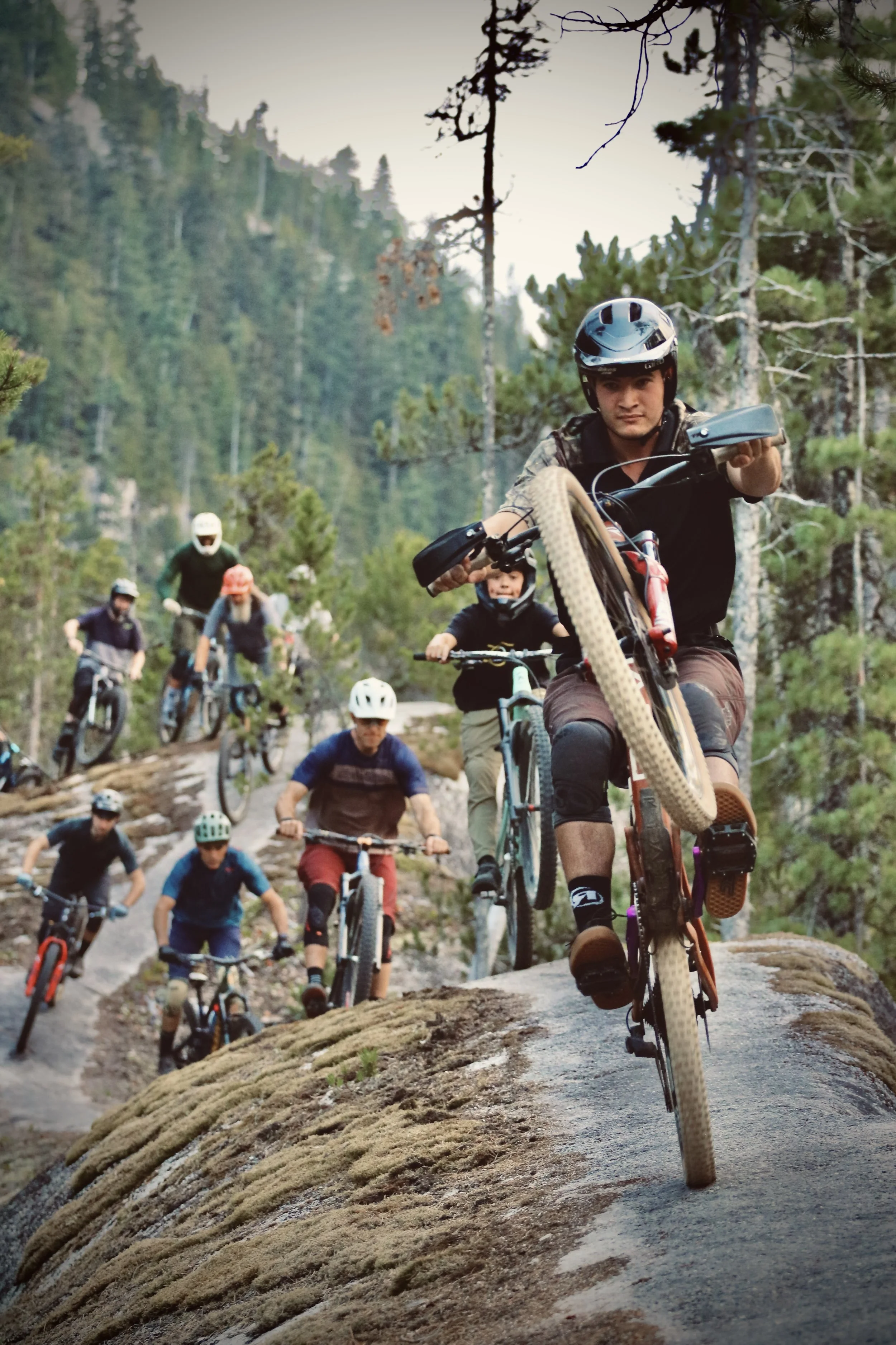 A group of people mountain biking on a rocky trail through a forested area with tall pine trees, all wearing helmets and protective gear, with the leading cyclist in focus.