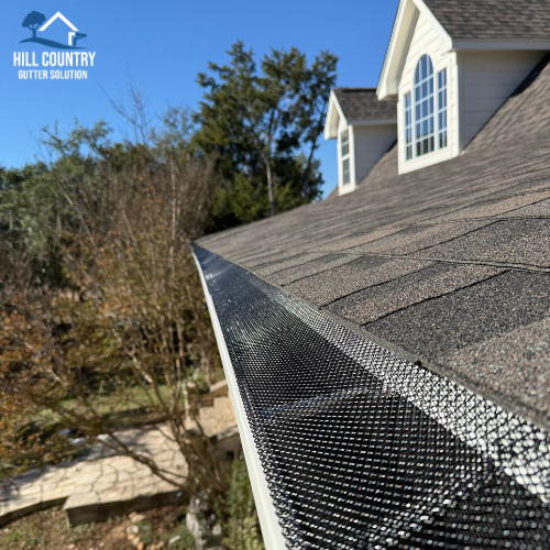 Close-up of roof with gutter guard installed along the edge, with a house featuring white siding and a gabled window in the background, and trees with some autumn foliage under a clear blue sky.