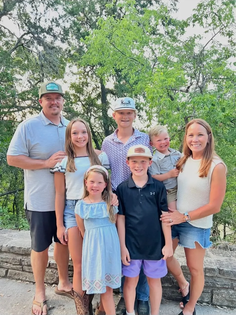 A happy family of eight posing outdoors on a stone wall surrounded by green trees, dressed casually for warm weather.