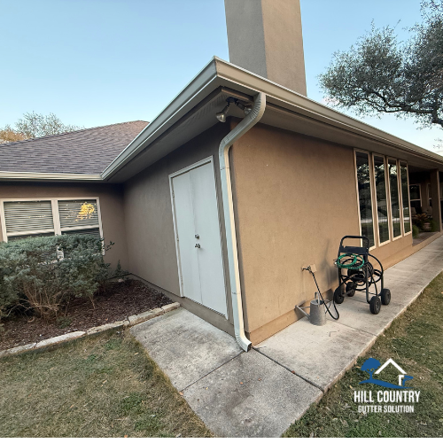 Backyard of a house with a beige exterior, a white door, large windows, a concrete walkway, and gutter system; gardening tools and a stroller near the wall.