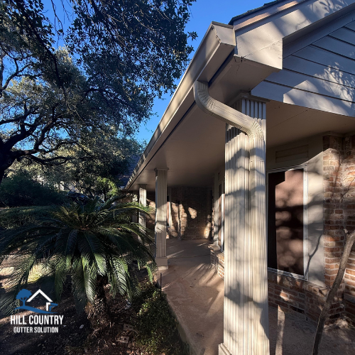 A house exterior with a porch supported by white columns, brick walls, a white gutter system, and surrounding trees.