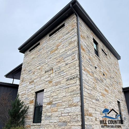 A stone building with a black roof and a downspout, featuring multiple small rectangular windows, and a logo for Hill Country Gutter Solution at the bottom right corner.