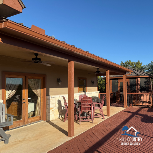 View of a covered patio with wooden support beams, a table with chairs, sliding glass doors, and a fenced porch area with a new gutter system, under a clear blue sky.