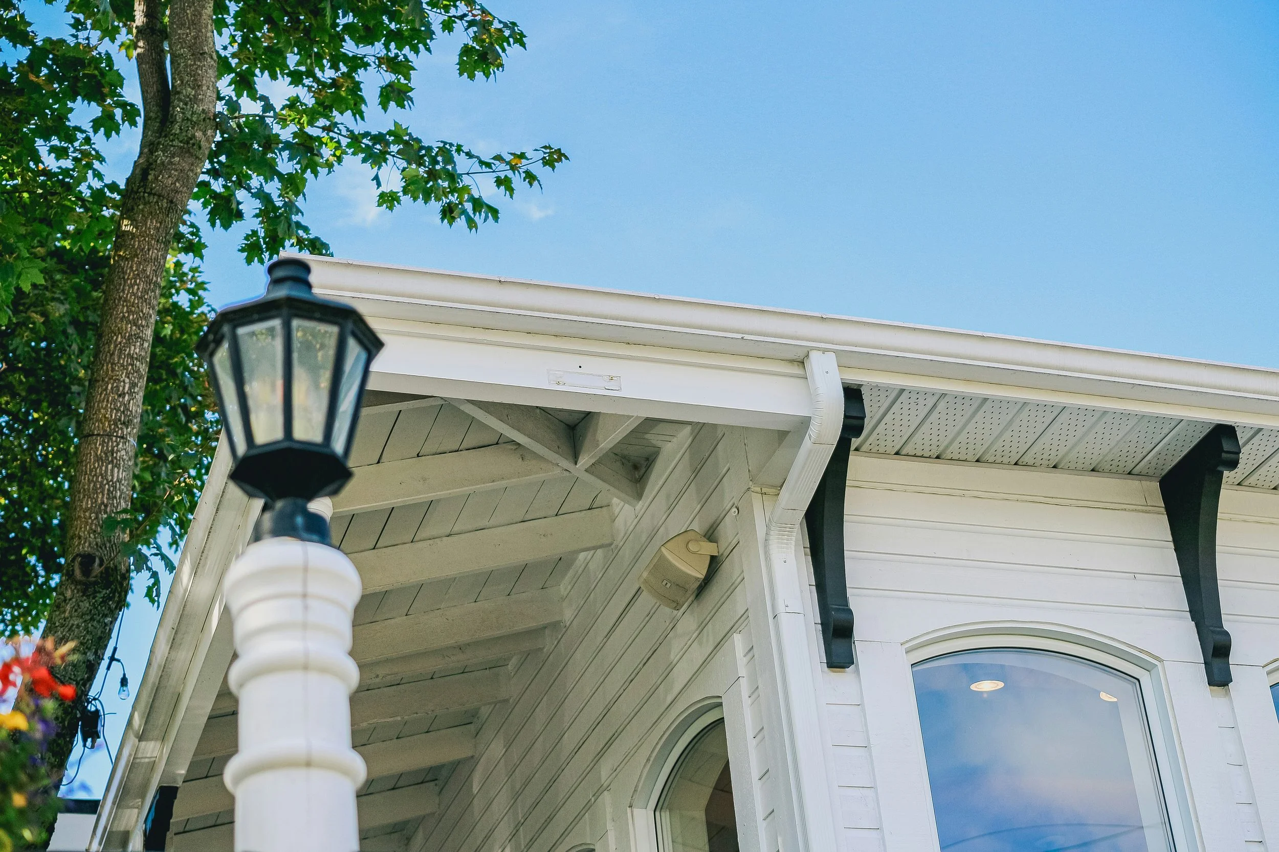 Close-up view of a white house corner with a window, black decorative brackets under the roof eaves, a white gutter, a white porch column, and a black lantern situated next to a maple tree with green leaves. The sky is clear and blue.