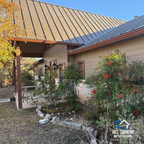Exterior of a house with a sloped metal roof, wooden siding, and a garden with red flowers and bushes.