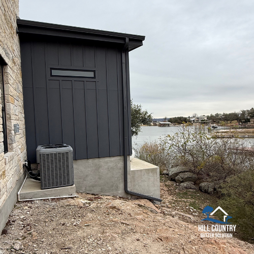 Exterior side of a modern house with dark gray siding, an air conditioning unit on a concrete pad, and a river or lake in the background with overcast sky.