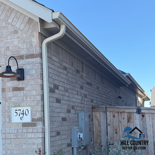 Close-up of a brick house wall with a black outdoor wall light, a white gutter pipe, electrical boxes, and a wooden fence with the Hill Country Gutter Solutions logo.