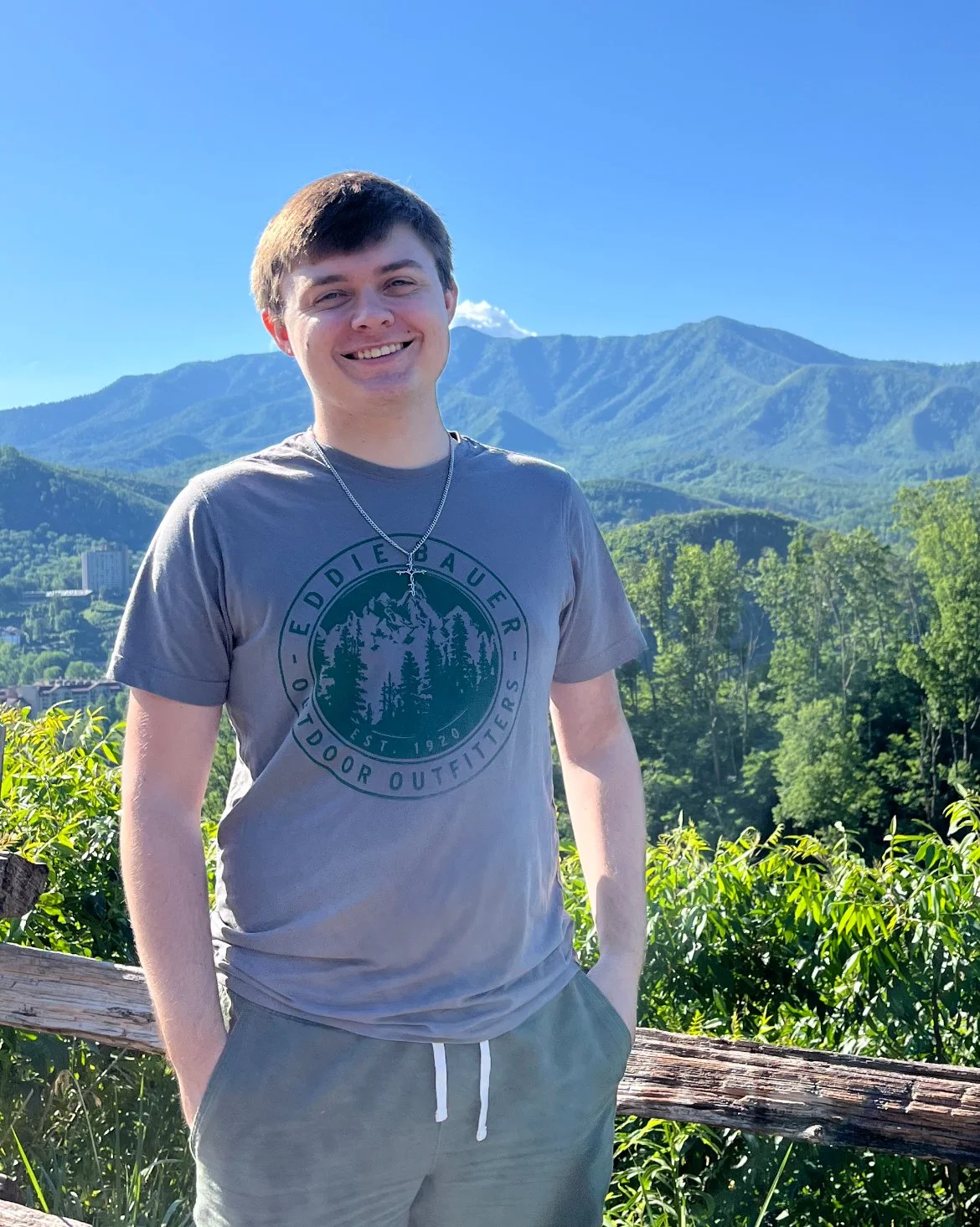 Young man smiling outdoors with mountains and green trees in the background on a sunny day.