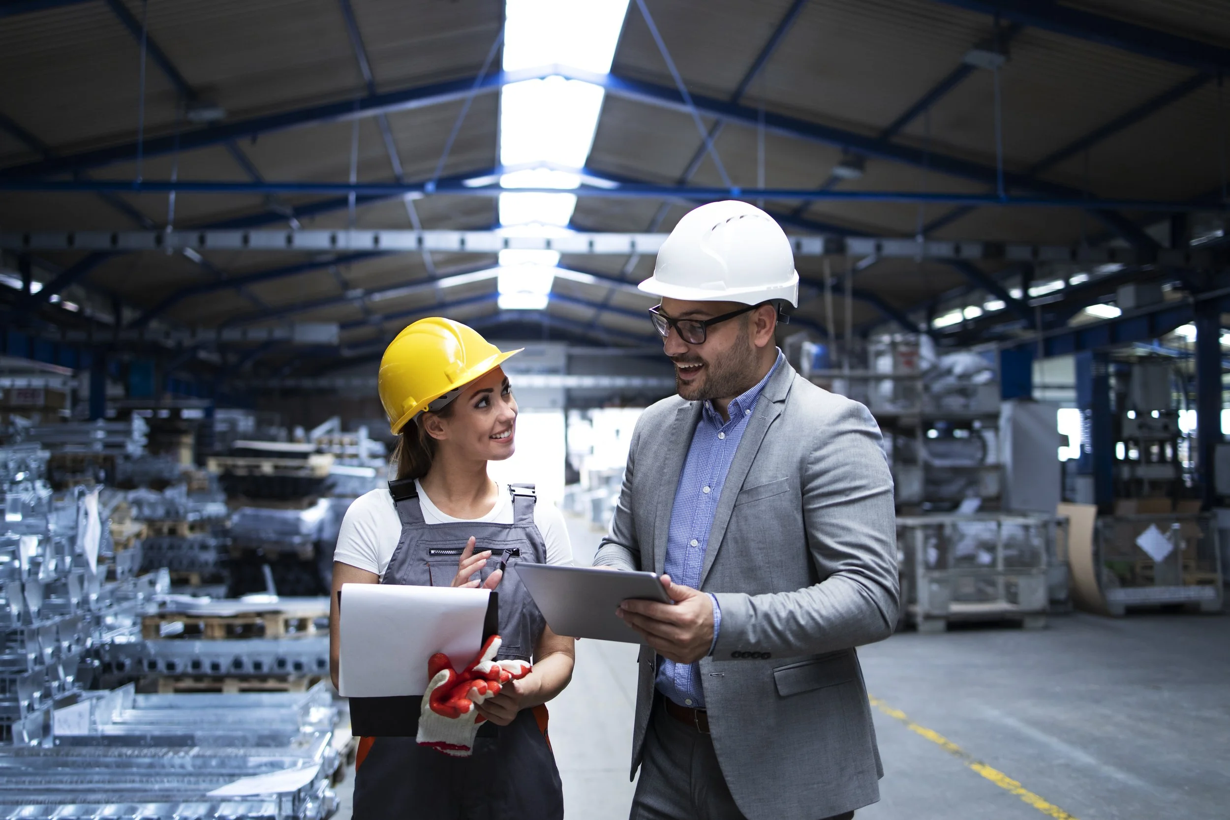 Two workers in safety helmets and work attire talking and smiling inside a warehouse with metal shelves and industrial equipment.