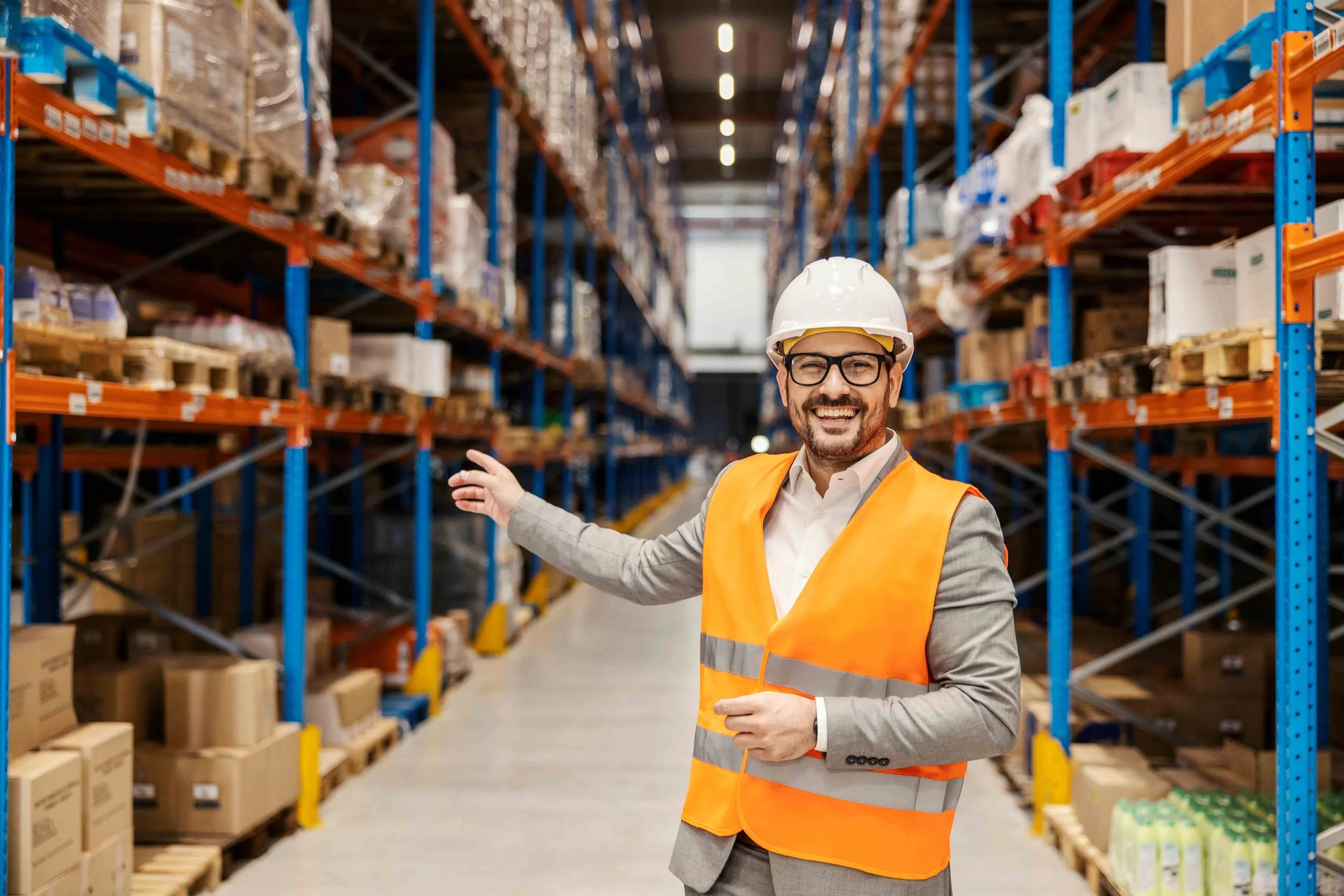 A man in safety glasses, a white hard hat, gray suit, and orange safety vest smiling and pointing in a warehouse aisle filled with shelves of boxes and products.