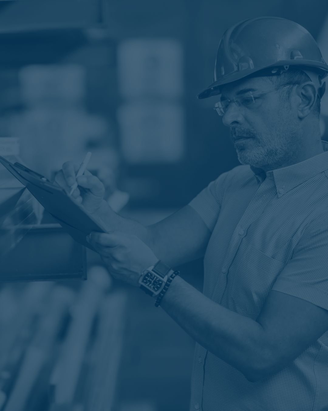 A man wearing a hard hat and safety glasses is looking at a clipboard, inspecting industrial equipment in what appears to be a factory or warehouse.