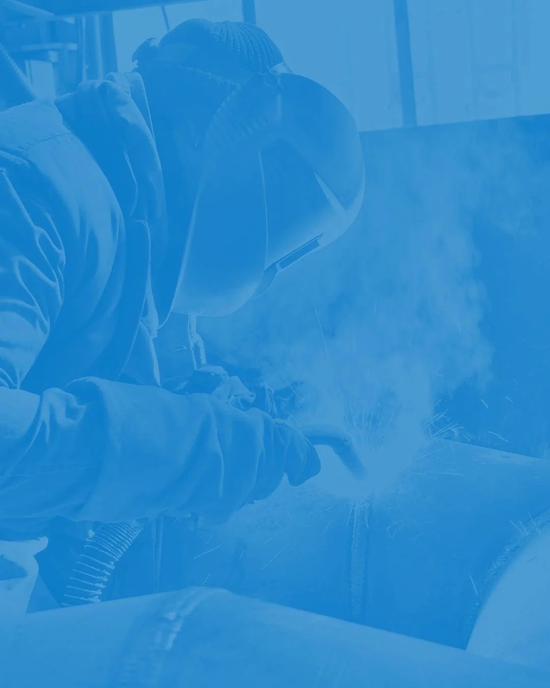 A person welding with sparks flying, wearing safety goggles, a cap, and a jacket in an industrial setting.