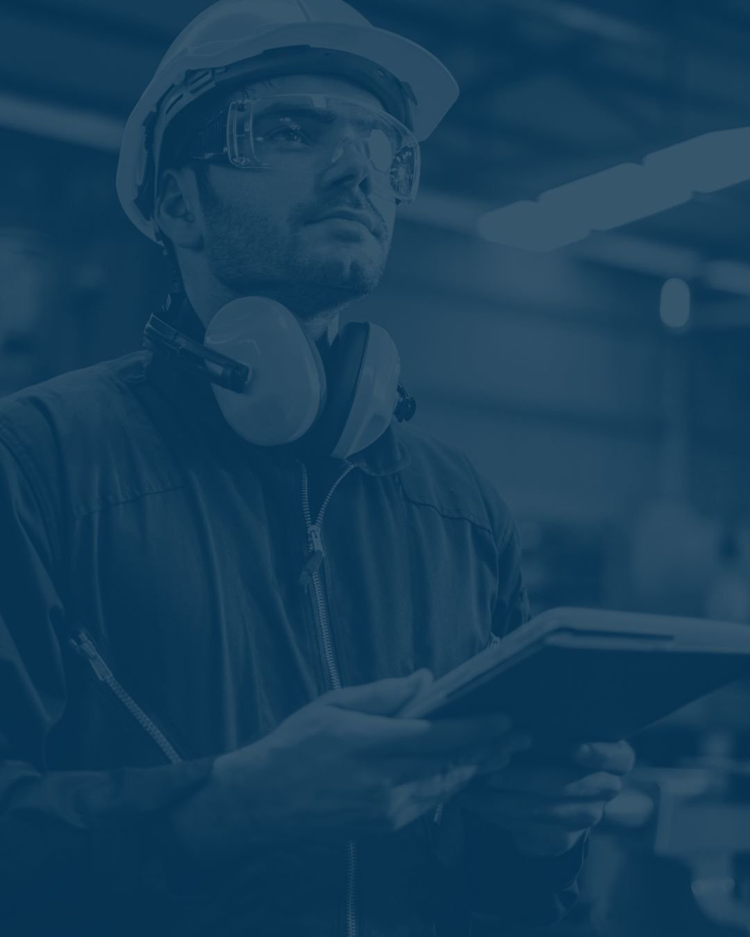 A man in protective gear, including a hard hat, goggles, a headset, and a leather jacket, holding a tablet or clipboard and working in an industrial or construction setting.
