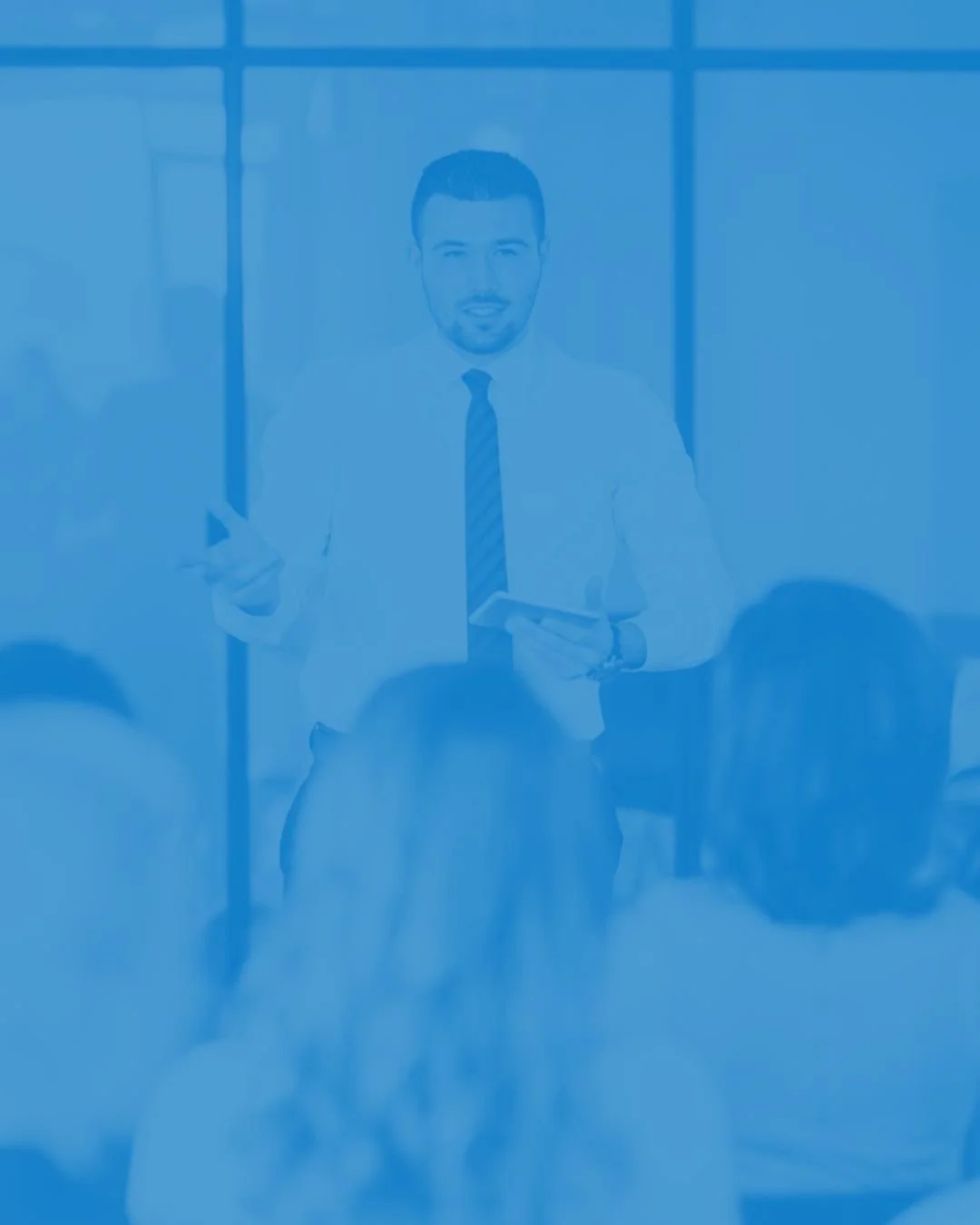 A man with a beard and dark hair, wearing a white shirt and striped tie, stands in front of a glass wall in a modern office setting, speaking to an audience.