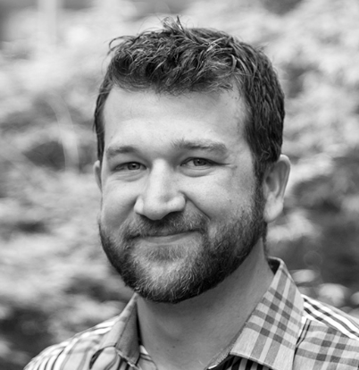 A man with a beard smiling outdoors, wearing a checkered shirt.