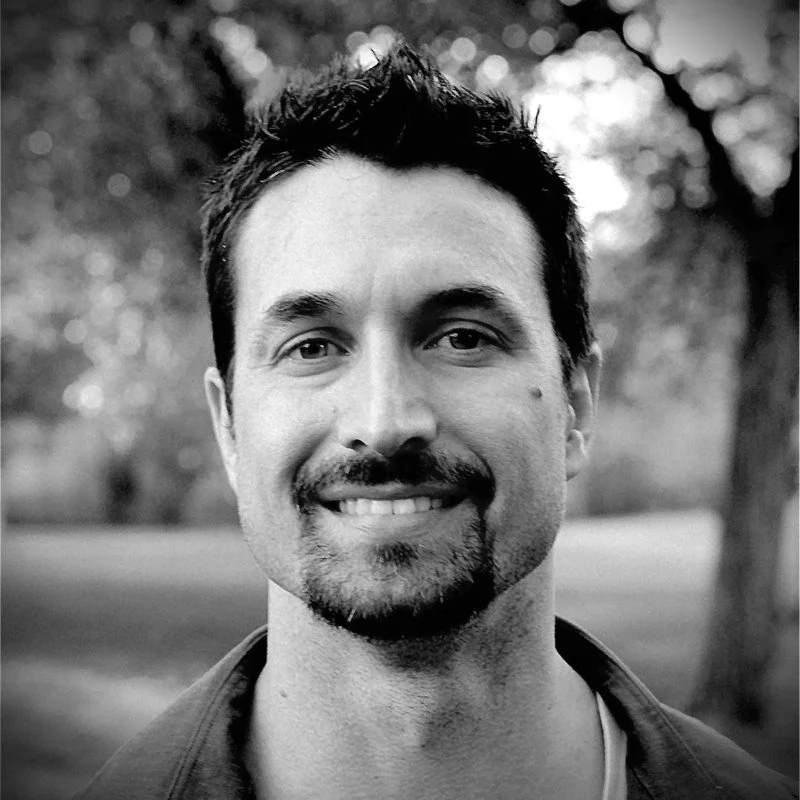 Black and white photo of a smiling man with dark hair and a goatee, outdoors with blurred trees in the background.