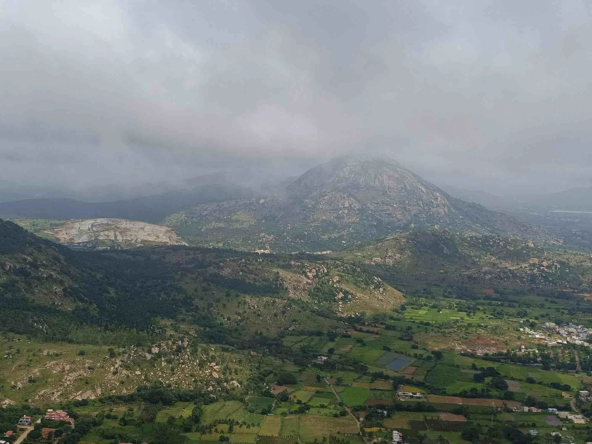 A mountainous landscape with a large hill or mountain partially covered by clouds, surrounded by green fields, some houses, and scattered trees in a rural area.
